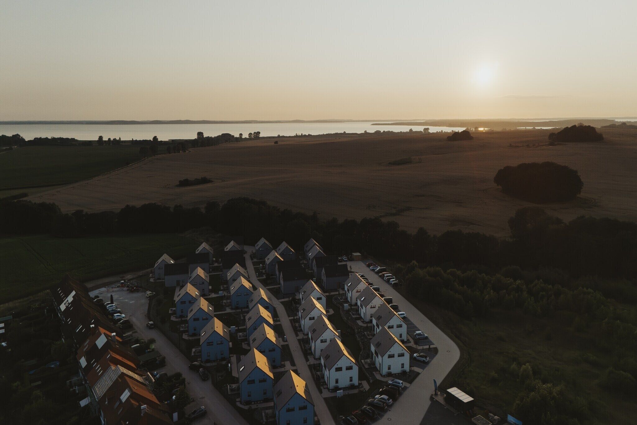 An aerial view of a residential area with houses arranged in rows, a road, and a body of water in the background.