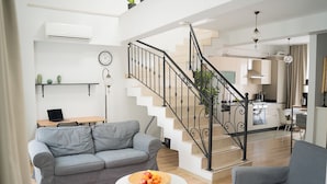 A modern living room with a grey sofa, a round coffee table with fruit, a staircase with a black railing, and a kitchen in the background. - Stylish Duplex with Private Terrace & Golden Horn View – 5 min to Istiklal (İstanbul)