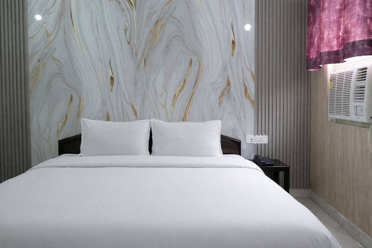 Una cama con ropa blanca, una pared con diseño de mármol, una mesita de noche con un teléfono y una ventana con cortina.