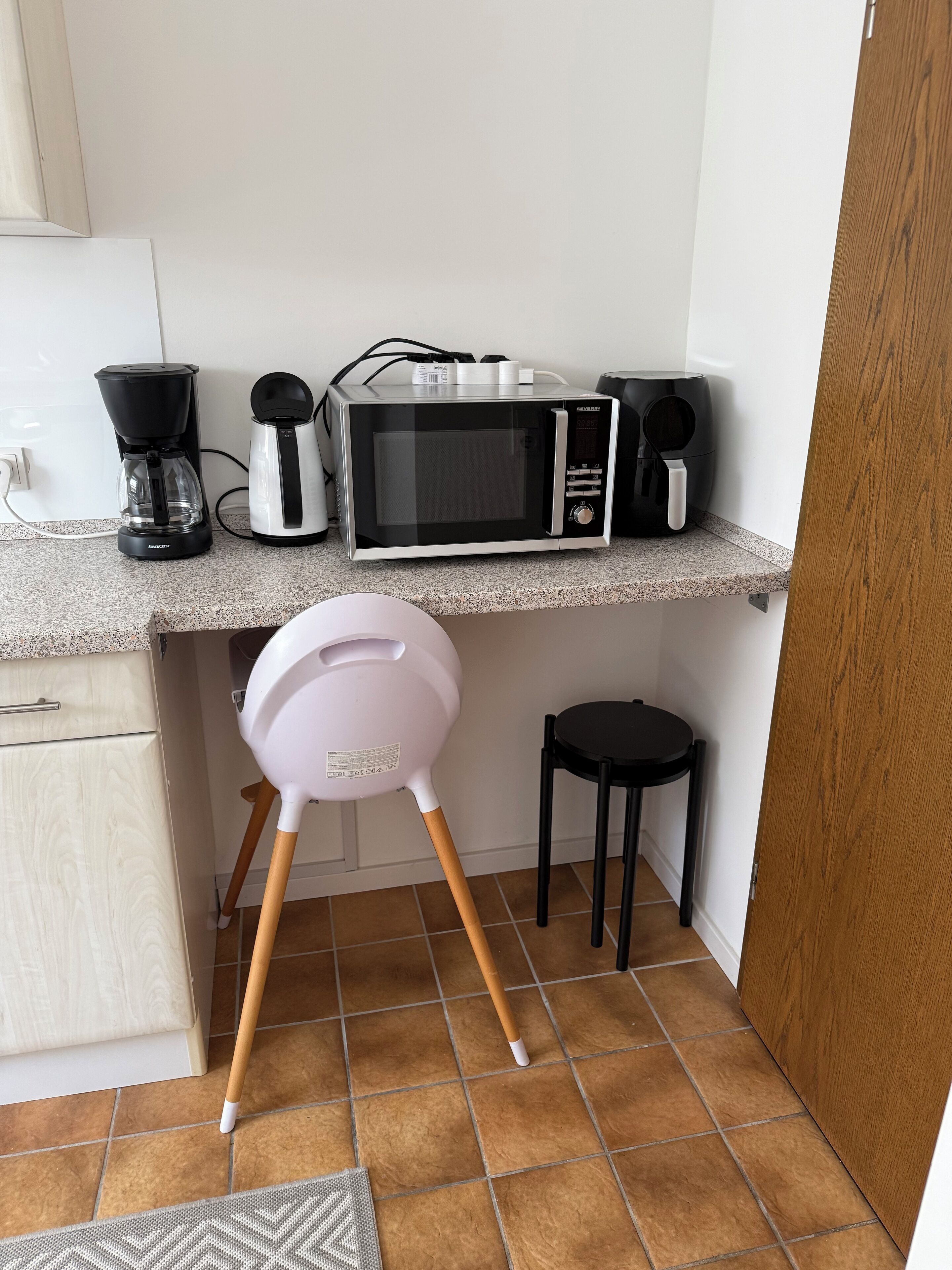 Una cocina con microondas, cafetera y un taburete pequeño.