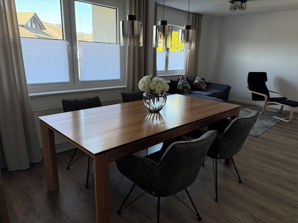Un comedor con una mesa de madera, sillas, un jarrón de flores y vistas al exterior a través de amplias ventanas.