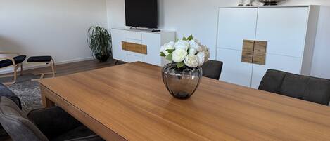 Un espace repas moderne avec une table en bois, des chaises à revêtement gris, un vase avec des fleurs blanches, une plante en pot et un meuble blanc sur lequel est posée une télévision.