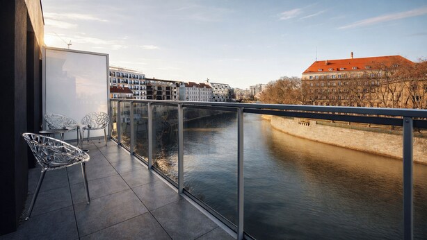 Ein Balkon mit Glasgeländer, der auf einen Fluss mit Blick auf Gebäude am anderen Ufer führt, mit Metallstühlen.