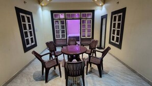 A room with a round table and chairs, a bed with a purple-lit window, and a door with a black frame. - Cozy Nirmala House Homestay in Varanasi (Varanasi)