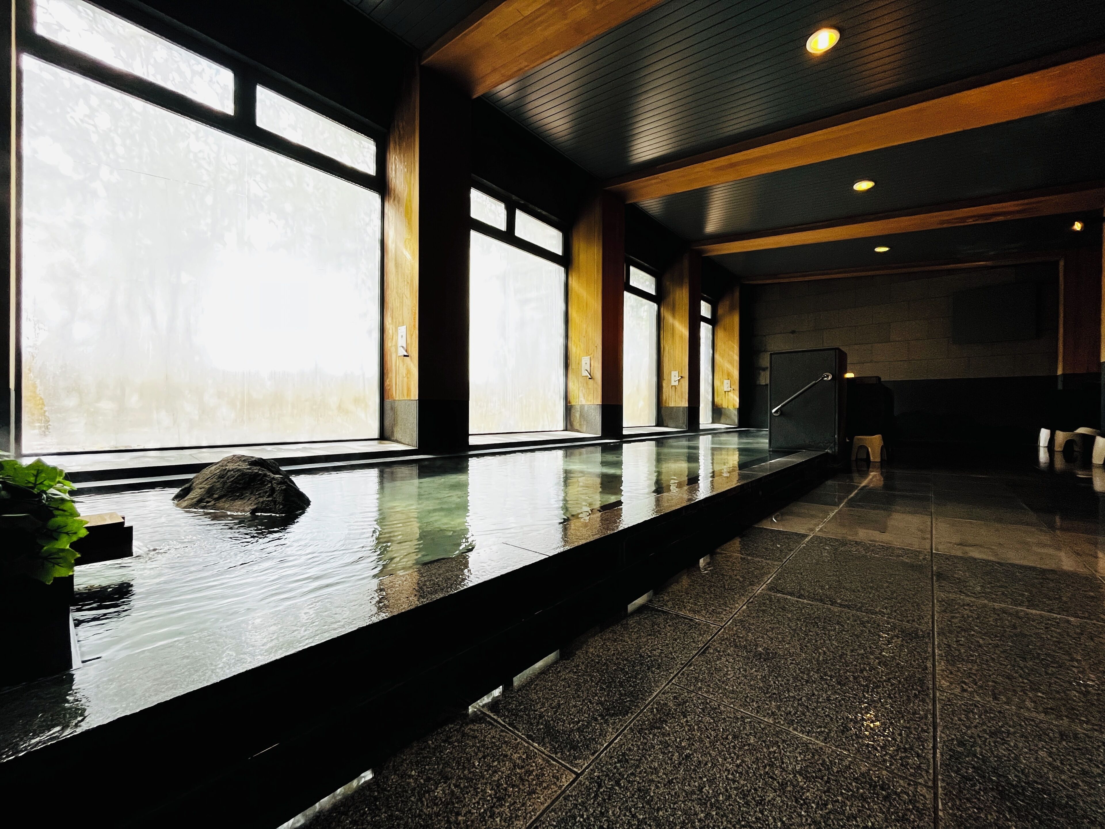 A modern indoor pool area with large windows, a stone floor, and a wooden ceiling.