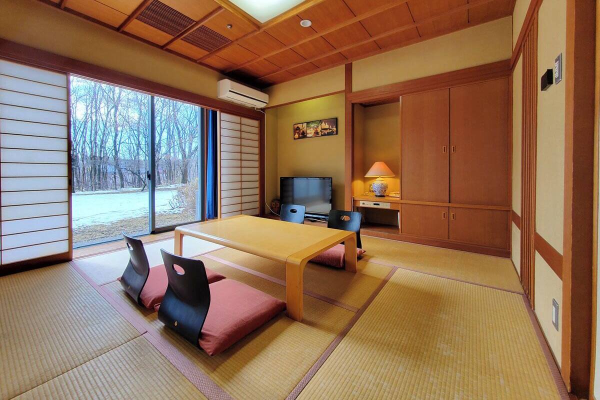 Une pièce de style japonais traditionnel, avec un sol recouvert de tatamis, une table basse, des sièges, un téléviseur et une porte coulissante donnant sur une vue sur les arbres.