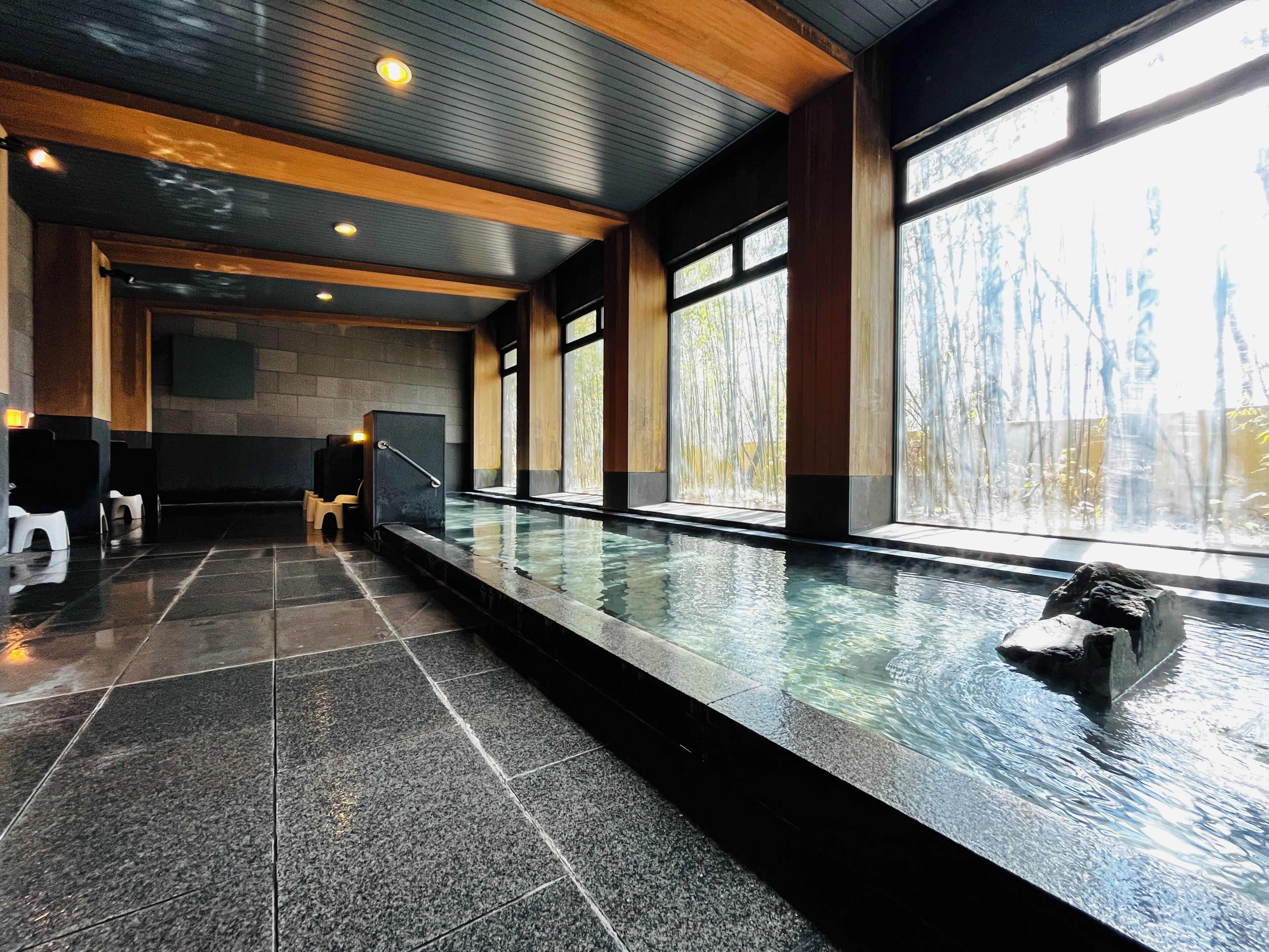 A modern indoor pool with clear glass walls, a stone floor, and wooden ceiling beams.