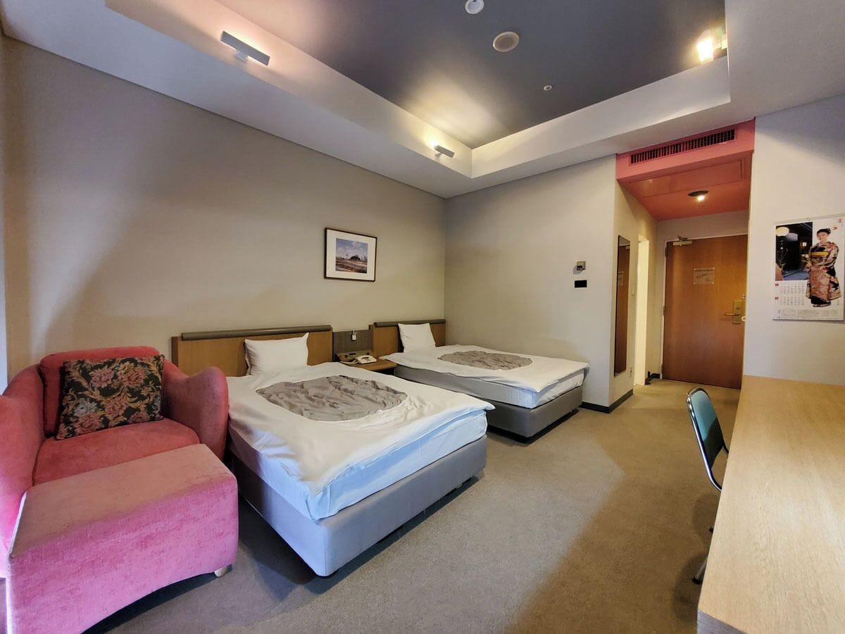 Habitación de hotel con dos camas, un sillón rosa, un escritorio y una puerta que da a otra habitación.