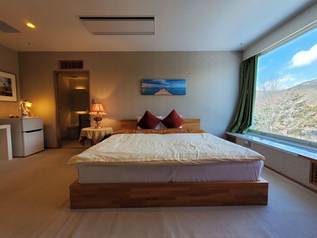 Quarto com cama, escrivaninha, cadeira, luminária, pintura e janela com vista.