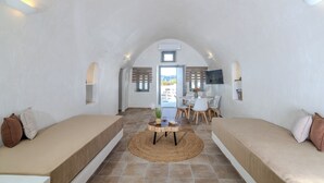 A room with a high arched ceiling, a central round table with a woven rug, two sofas, a dining area with chairs, and a TV mounted on the wall.