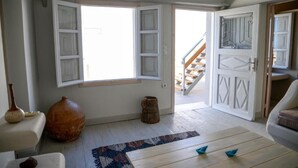Una habitación con un ventanal grande, piso de madera, puerta blanca con paneles decorativos y una pequeña mesa con un objeto azul encima.