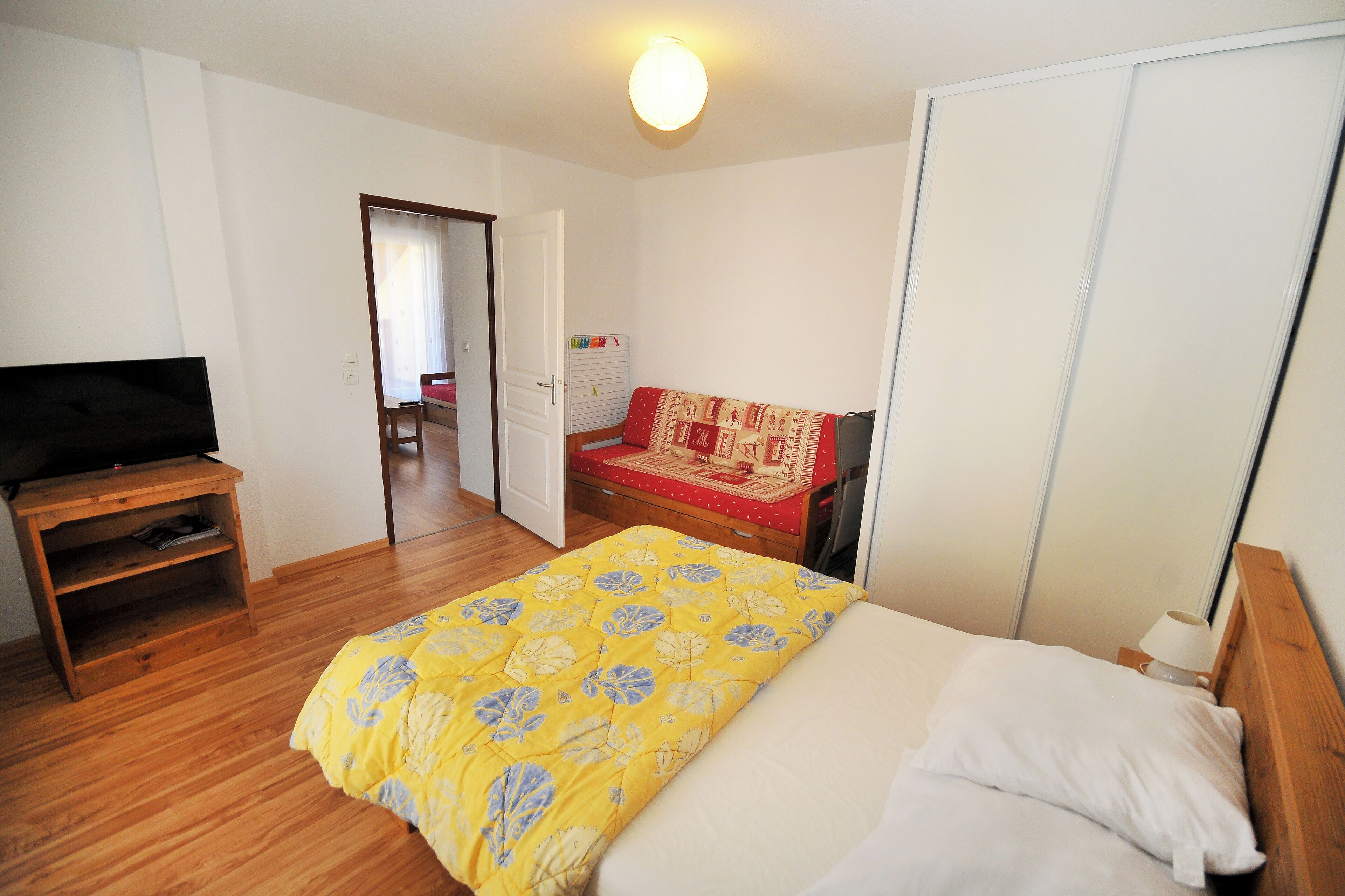 A bedroom with a bed, a TV on a stand, a wardrobe, and a door leading to another room.