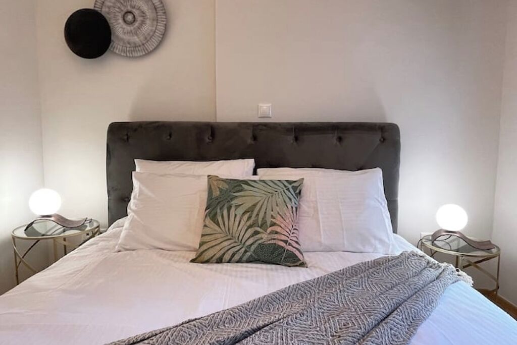Een bed met een gestoffeerd hoofdeinde, wit beddengoed, een geborduurde plaid en twee bedlampjes.