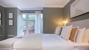 A bedroom with a bed, a bedside table, a window with curtains, and a wall-mounted light fixture. - The Abbey Mews House (London)
