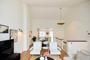 Een moderne woonkamer met een zwart tv-meubel, witte stoelen, een eettafel en een trap naar een hoger niveau.