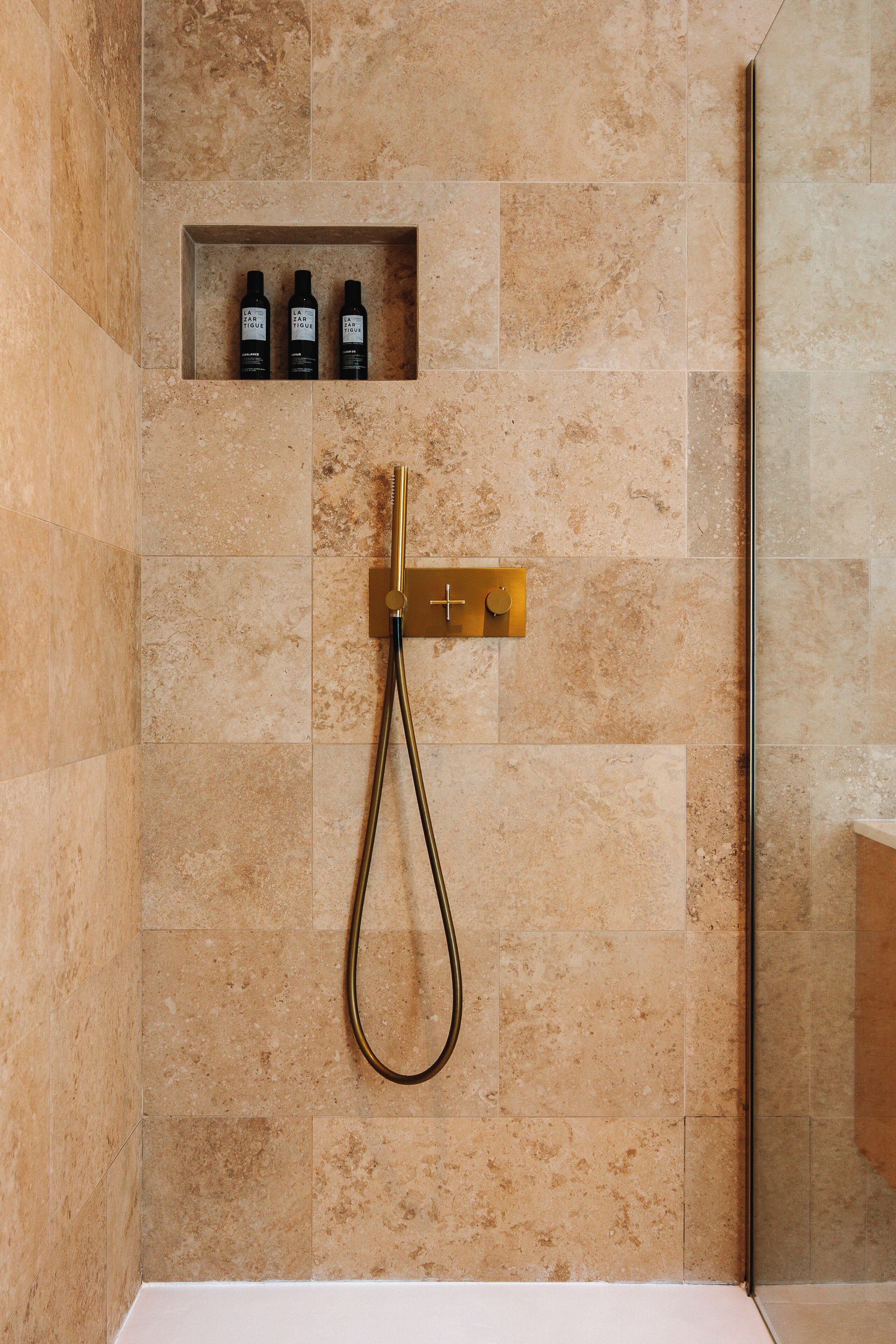 A shower with a gold showerhead and control panel, beige tiles, and a shelf with three bottles.