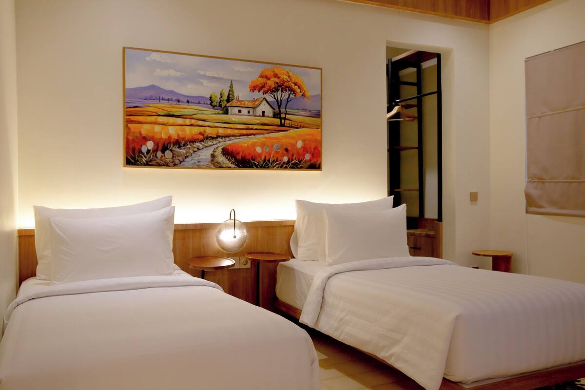 Een hotelkamer met twee bedden, een schilderij van een landschap, een nachtkastje met een lamp en een houten hoofdeinde.