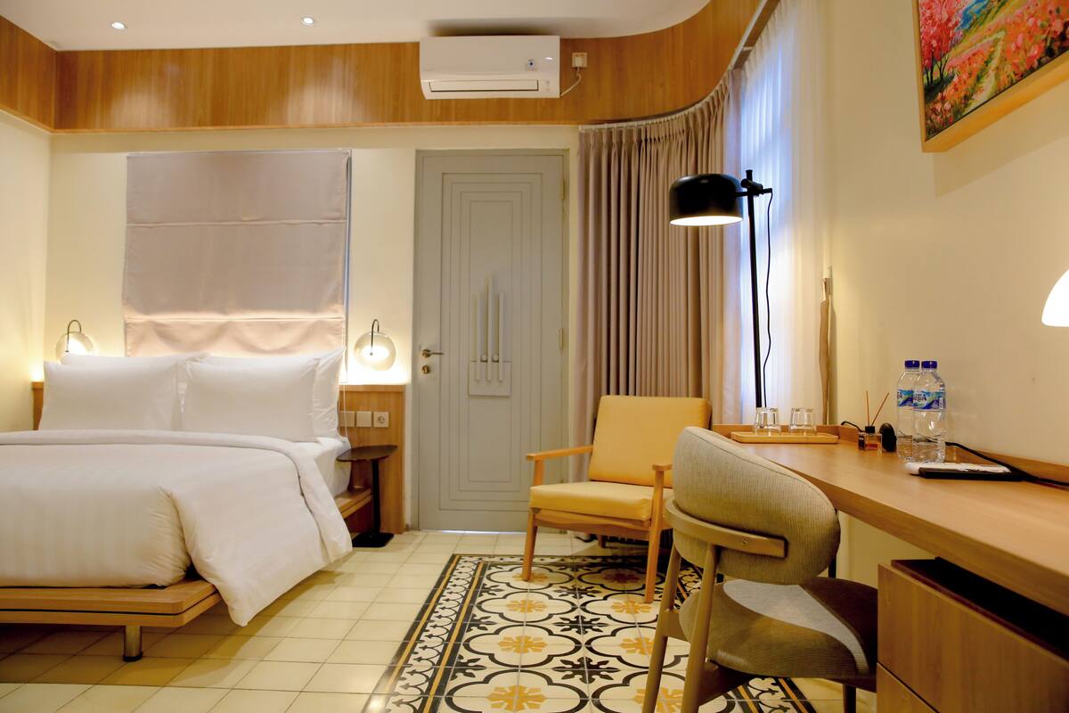 Een hotelkamer met een bed, een bureau, een stoel, een lamp, een waterfles en een schilderij aan de muur.