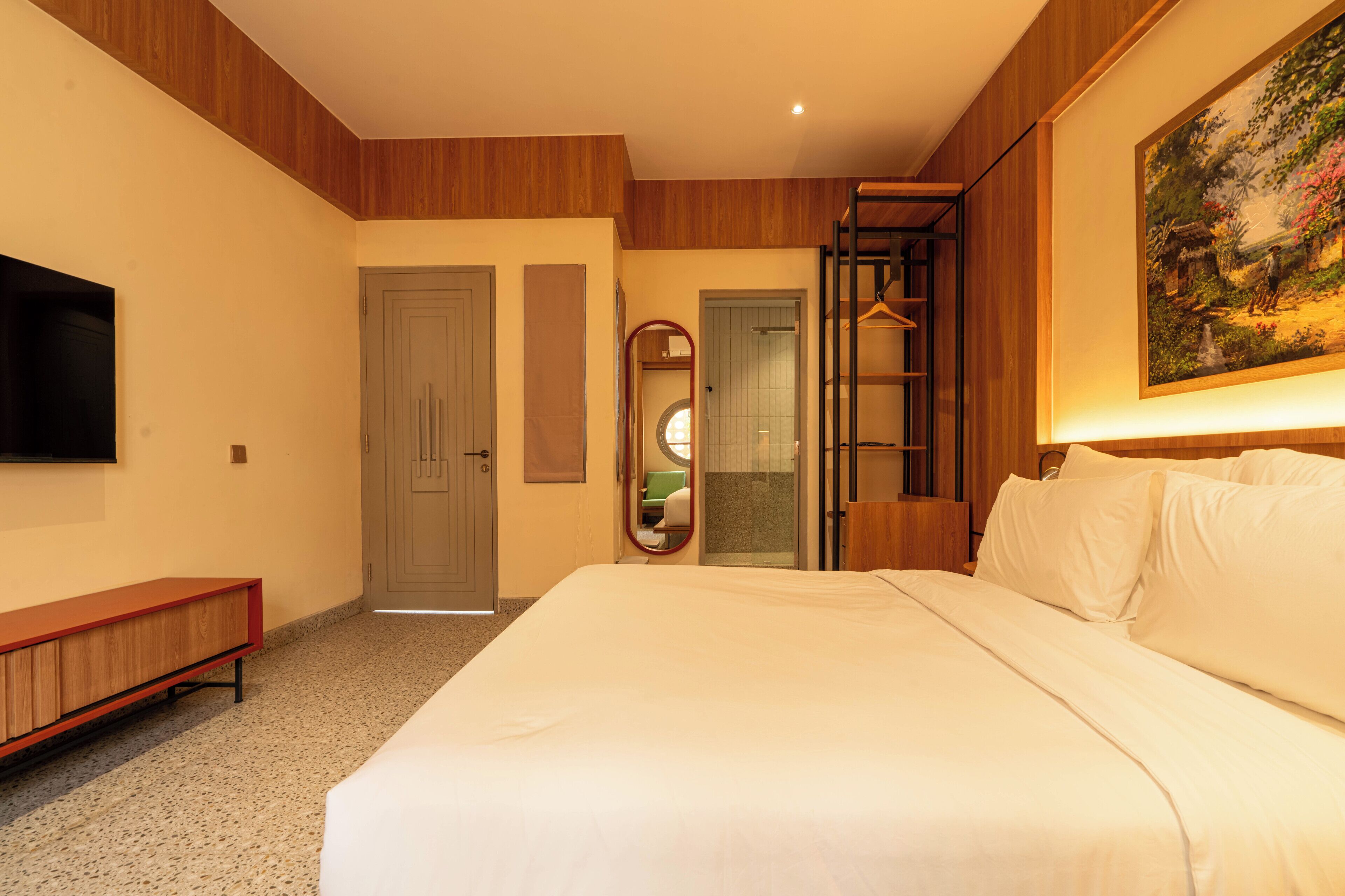 A hotel room with a bed, a TV, a bench, a mirror, and a painting on the wall.