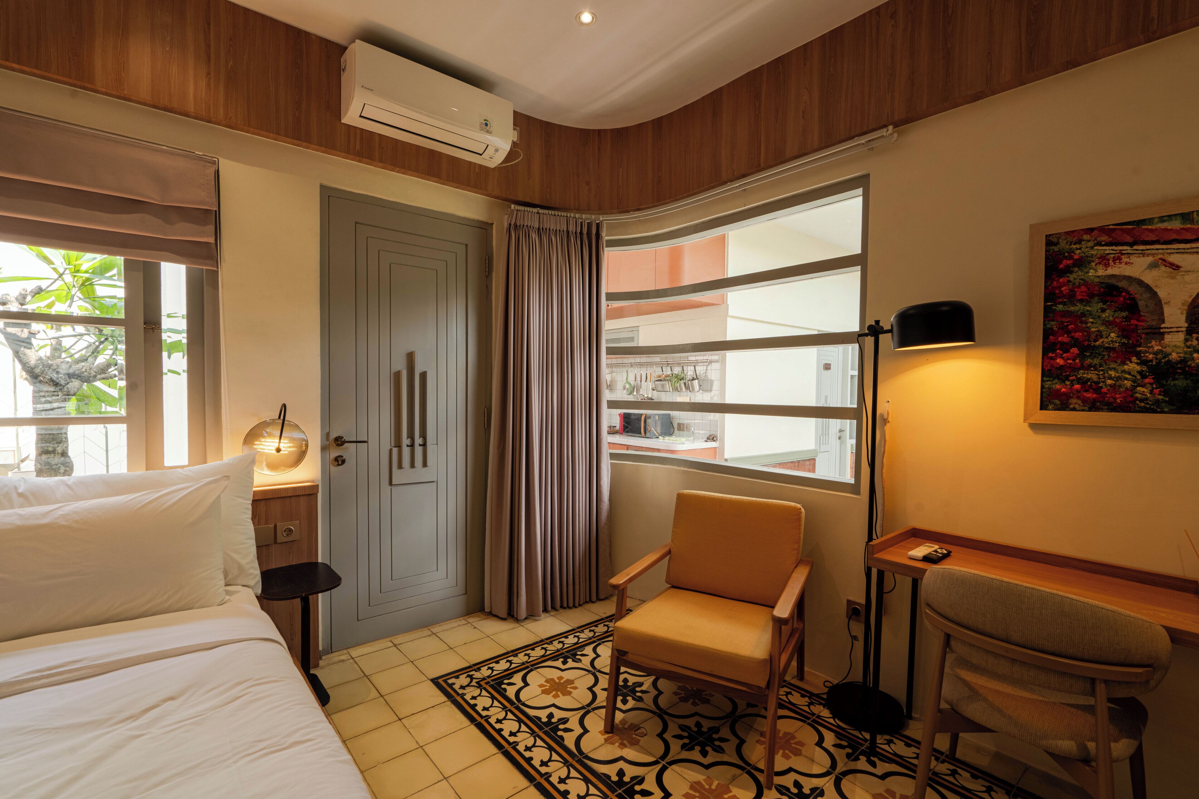 A hotel room with a bed, a chair, a desk, a window with curtains, and an air conditioning unit.