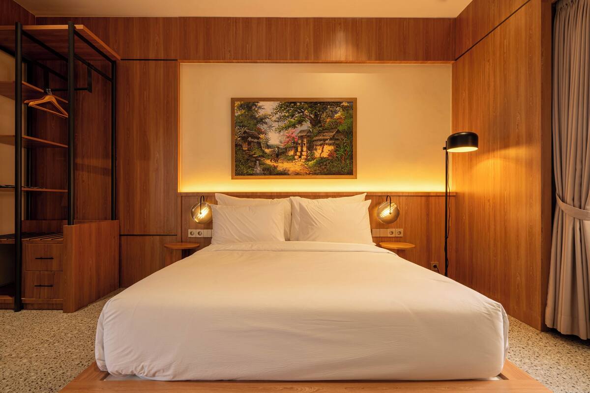 Een netjes opgemaakt bed met hoofdbord, een houten garderobe en een schilderij aan de muur.