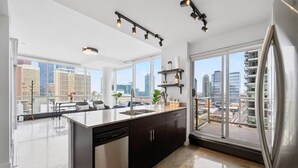 Cozinha moderna com uma ilha grande, armários pretos e uma geladeira de aço inoxidável. Várias janelas oferecem vista para a paisagem urbana.