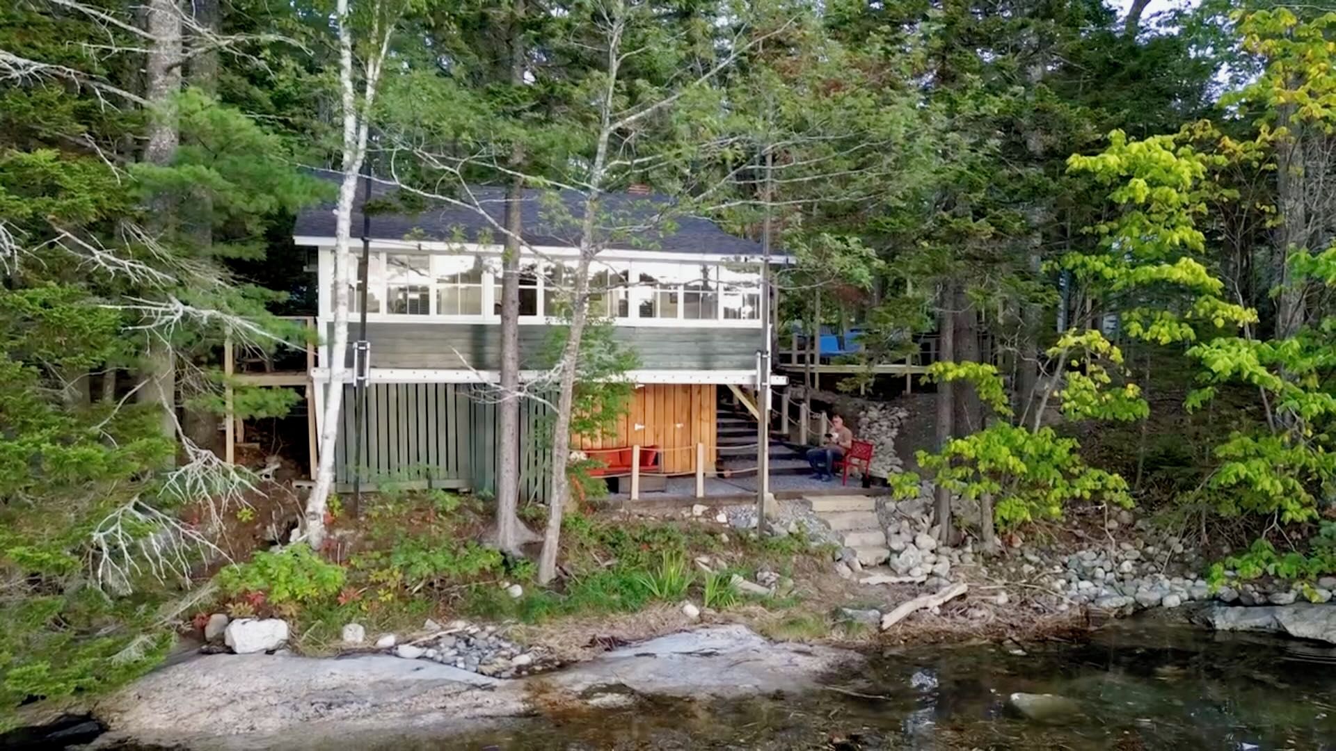 Una cabaña de dos pisos, rodeada de un bosque denso, cerca de un arroyo pequeño.