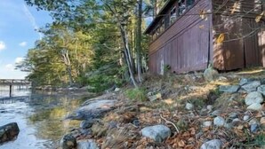 A riverside cabin with a wooden deck, surrounded by trees and a rocky shoreline.