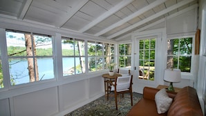 A sunroom with large windows, a brown leather sofa, a wooden chair, and a small table with a lamp.