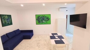 A room with a blue sofa, a white table with black placemats, a flat-screen TV, and two abstract green paintings on the walls. - Prince Apartment (Napoli)