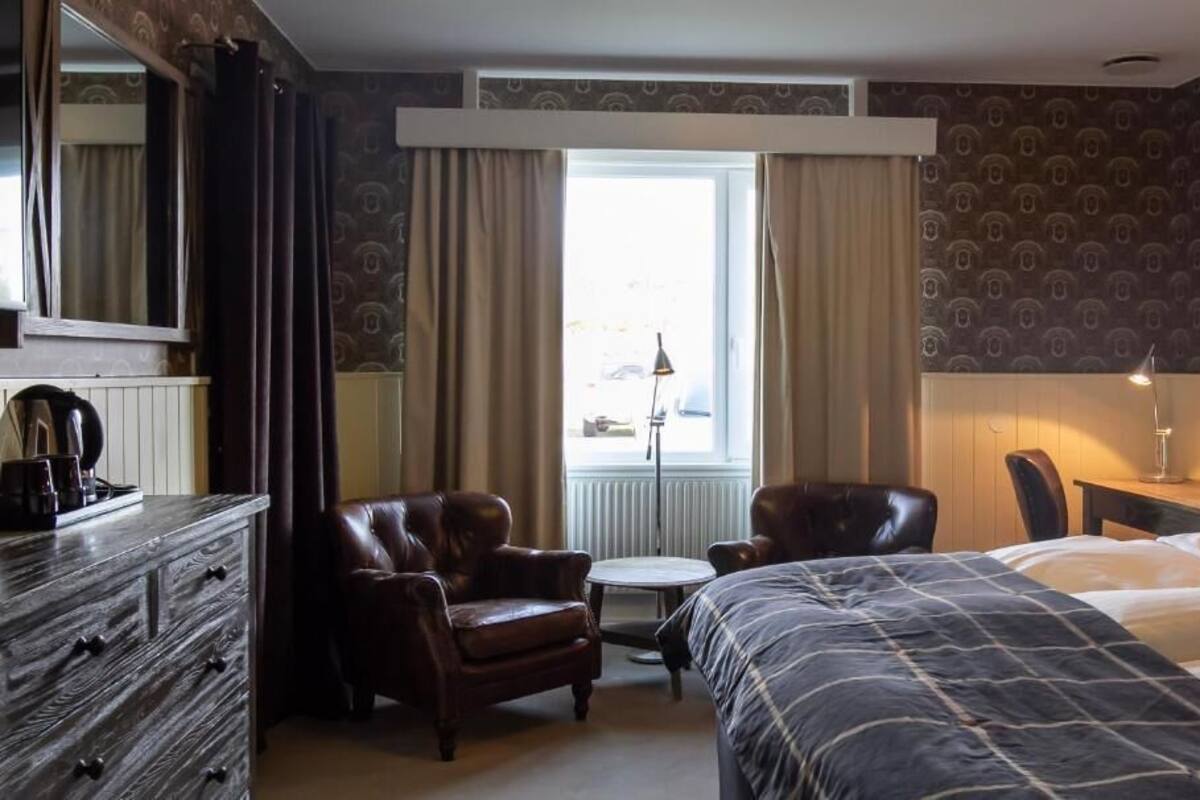 Hotelkamer met een bed, twee stoelen, een kleine tafel, een dressoir, een raam met gordijnen en een radiator.