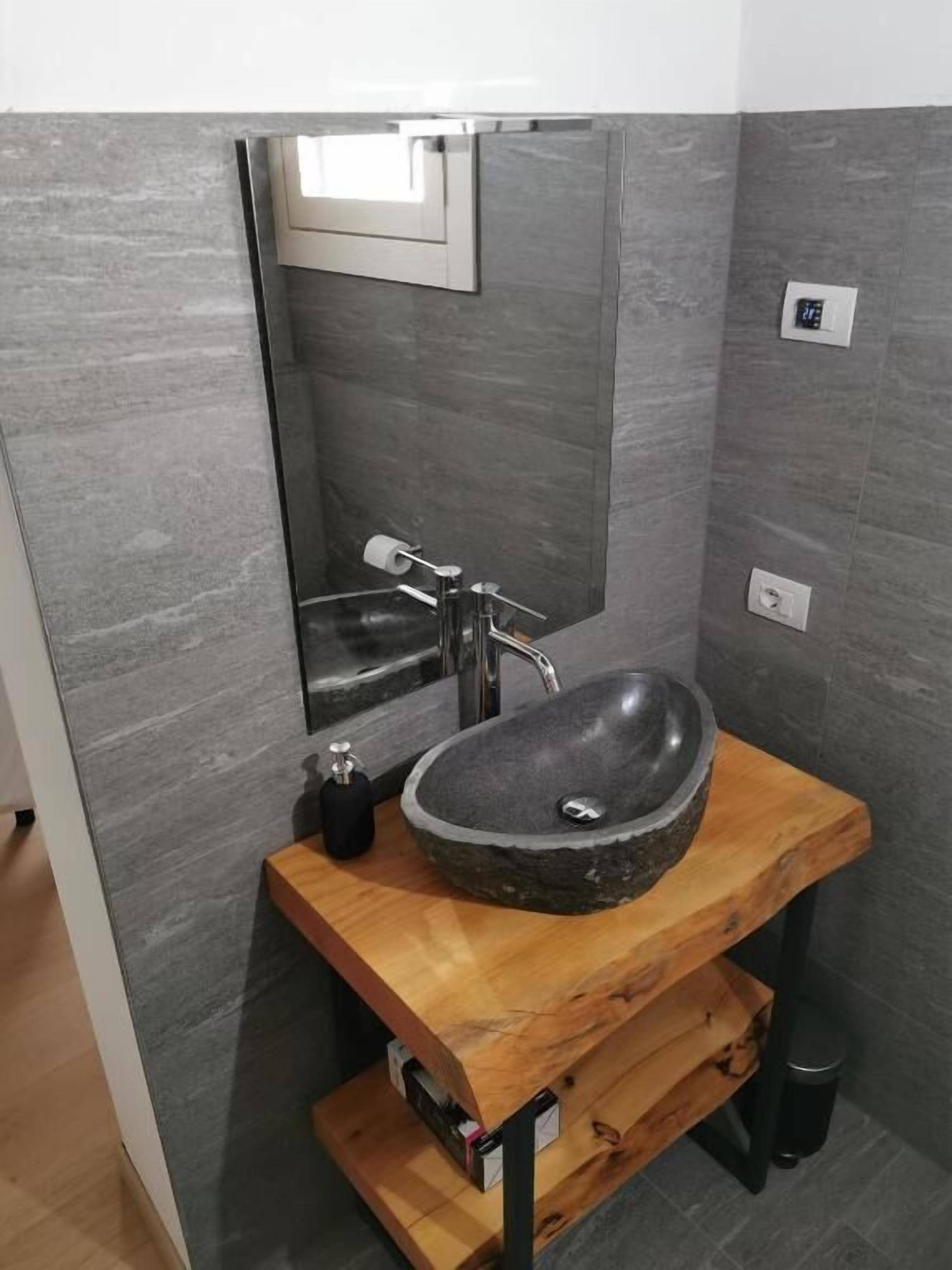 A bathroom with a stone sink, a mirror, a faucet, a soap dispenser, and a trash bin.