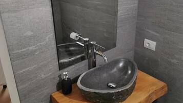 A bathroom with a stone sink, a mirror, a faucet, a soap dispenser, and a trash bin.