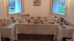 Un tavolo per la colazione a buffet con diversi cibi e condimenti, due finestre con vista sul verde e un quadro appeso al muro.