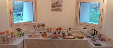 Une table de buffet proposant divers aliments et accompagnements pour le petit-déjeuner, deux fenêtres donnant sur un paysage verdoyant et un tableau encadré accroché au mur.