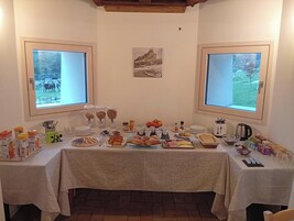 Una mesa de desayuno buffet con diversos alimentos y aderezos, dos ventanas con vistas a zonas verdes y un cuadro en la pared.
