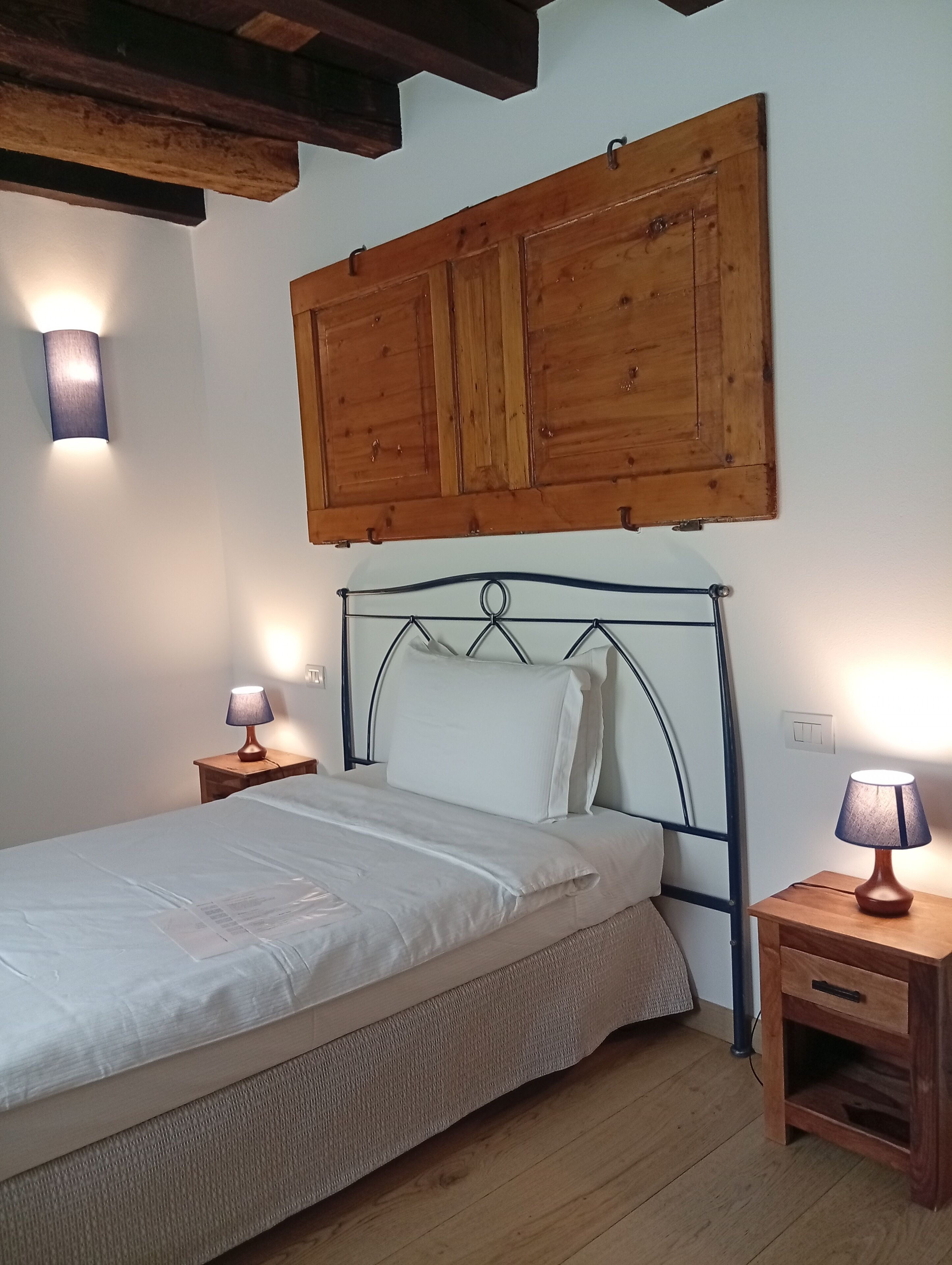 Un dormitorio con cama, cabecera de madera, dos mesitas de noche con lámparas y un armario de madera sobre la cama.
