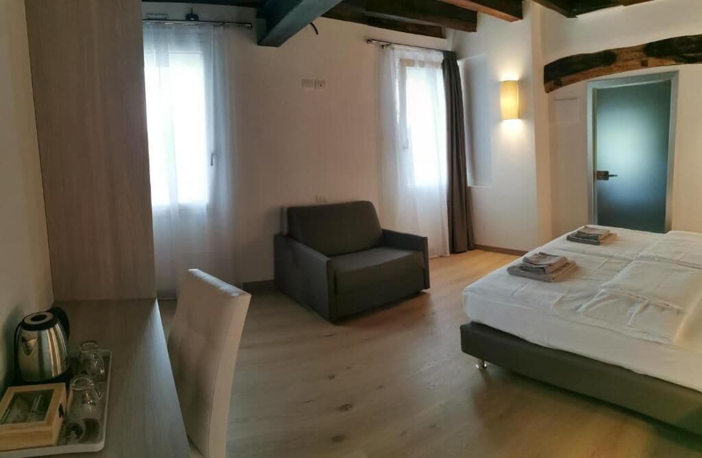 Una habitación con una cama, una silla, un escritorio con hervidor y vasos, y una puerta con luz.