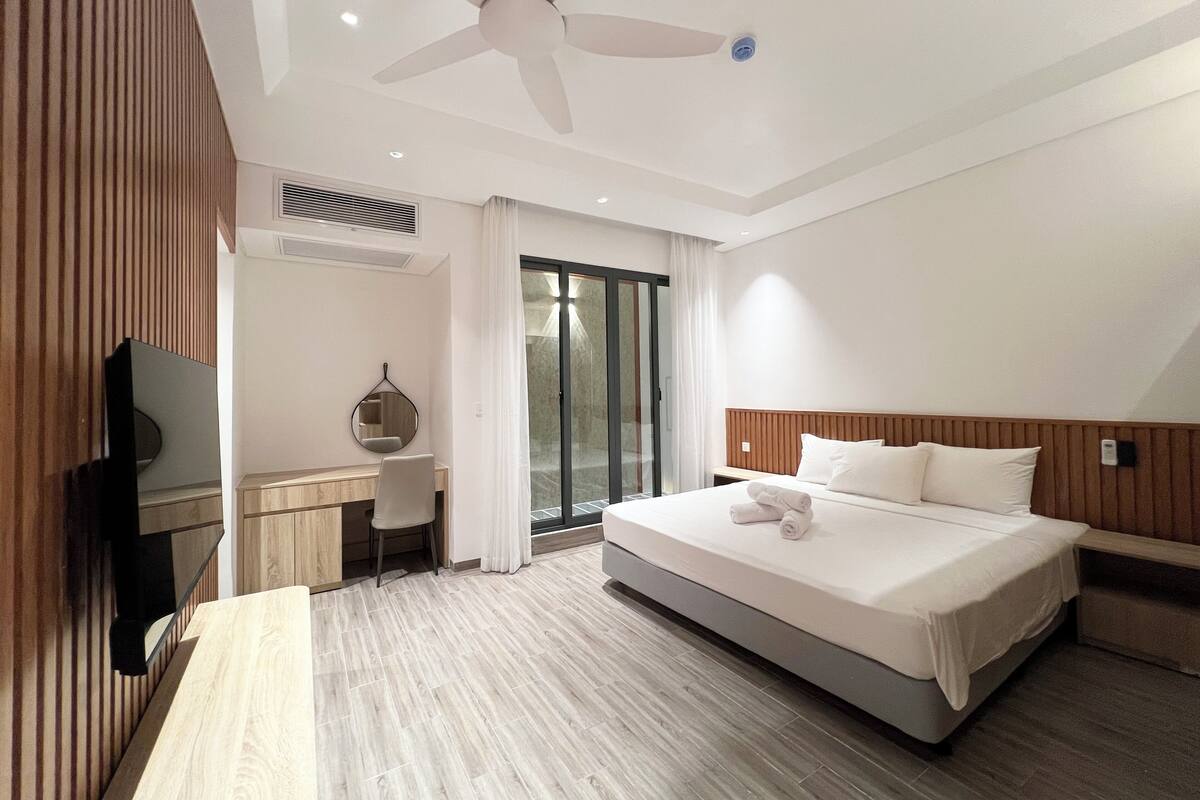 A hotel room with a bed, a desk, a chair, a television, a ceiling fan, and a sliding glass door.