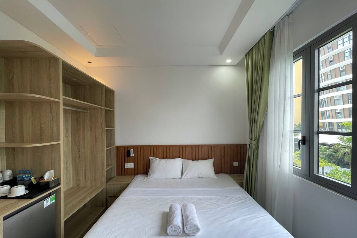 A hotel room with a bed, a wooden wardrobe, a small table with cups and a bottle, and a window with curtains.