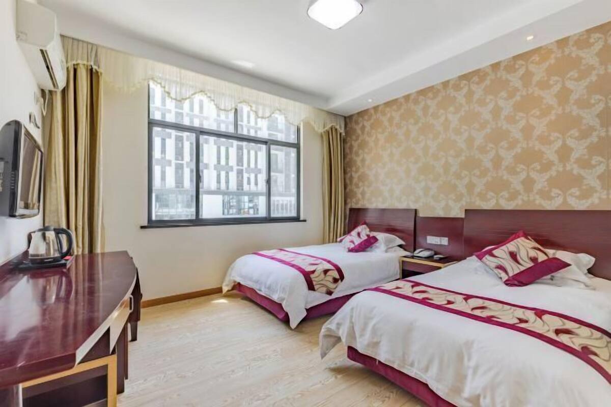 A hotel room with two beds, a desk, a window with curtains, and a patterned wall.