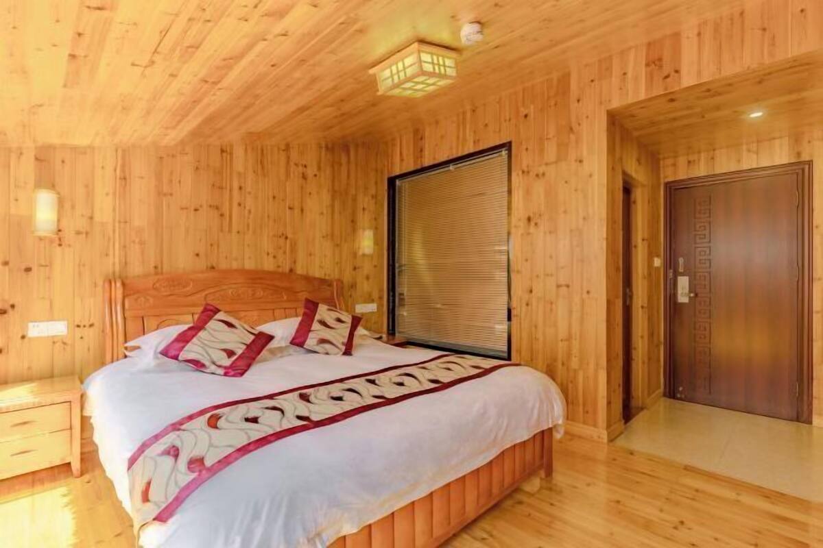 A wooden-paneled room with a bed, a window with blinds, a door, and a small table.