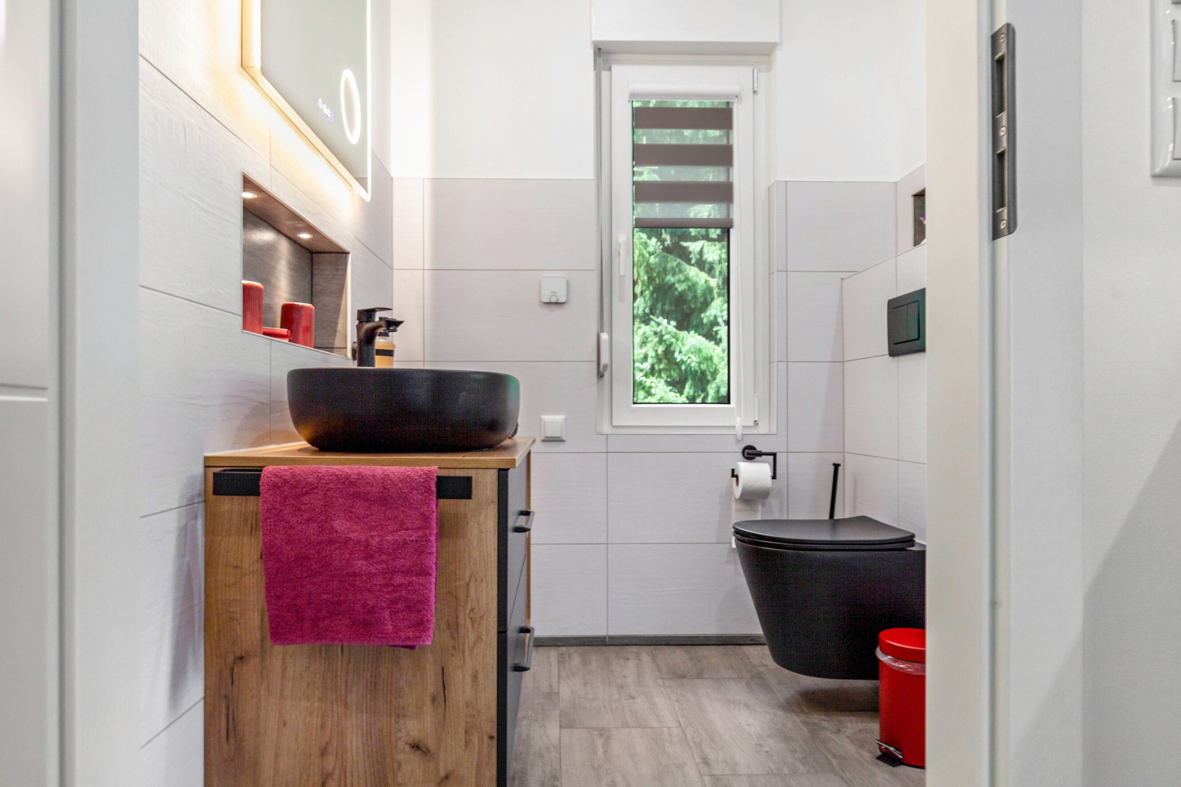 Un baño moderno con tocador de madera, un lavamanos grande, una bañera negra y una ventana con vistas al verdor.