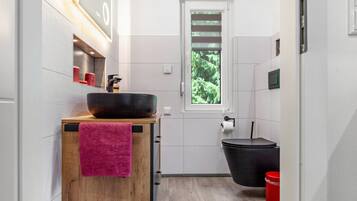 Un baño moderno con tocador de madera, un lavamanos grande, una bañera negra y una ventana con vistas al verdor.