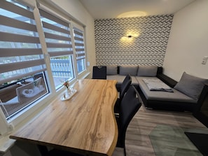 Una sala de estar moderna con una mesa de comedor de madera, un sofá gris con cojines, una pared con diseño y una ventana con persianas.