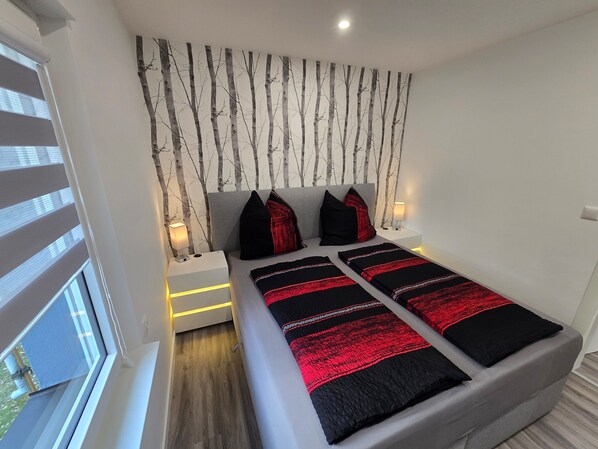 Un dormitorio con cama, dos mesitas de noche, una lámpara de pared y un ventanal con persianas.