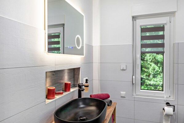 Ein modernes Badezimmer mit einem großen, schwarzen Waschbecken, einem Spiegel mit integrierter Beleuchtung, einem Fenster mit Jalousien und einer kleinen Ablage mit Pflegeprodukten.
