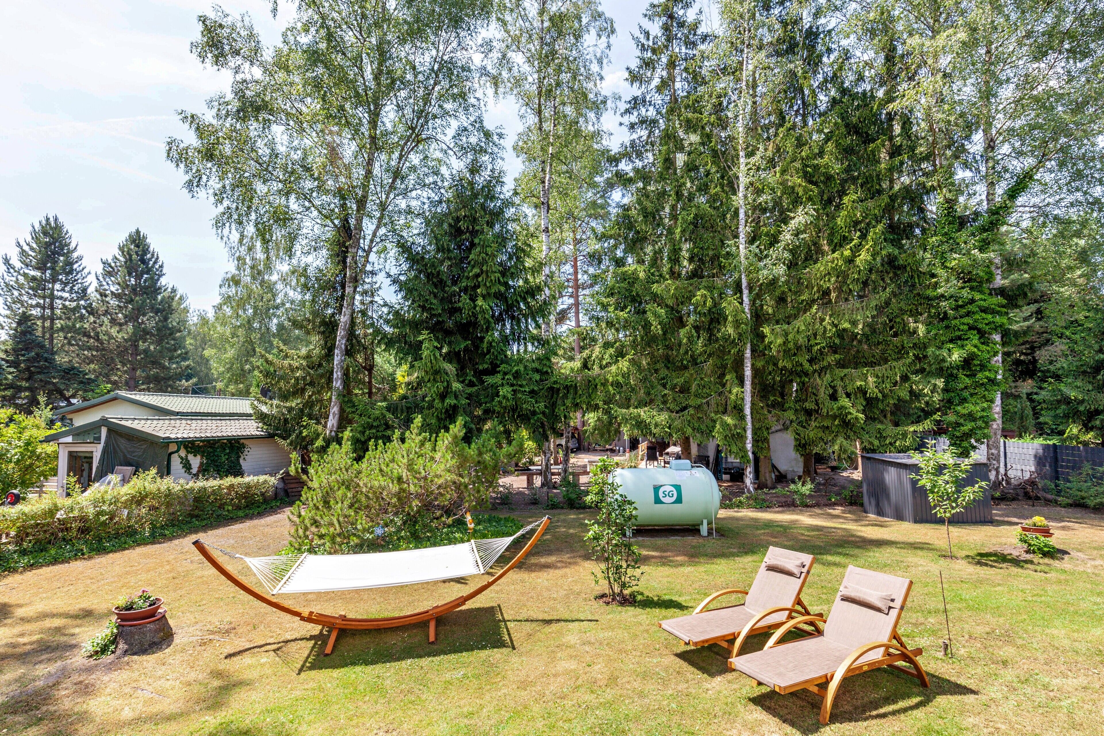 A hammock, two lounge chairs, a small shed, and a large water tank in a backyard with tall trees.