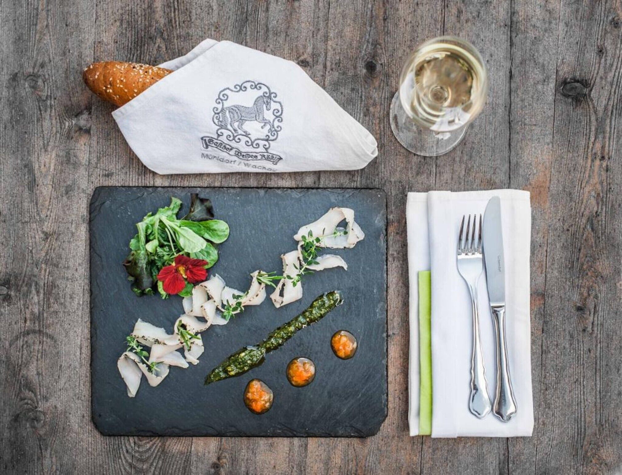 A slate serving board with food, a napkin, cutlery, a glass of white wine, and a bread roll on a wooden table.