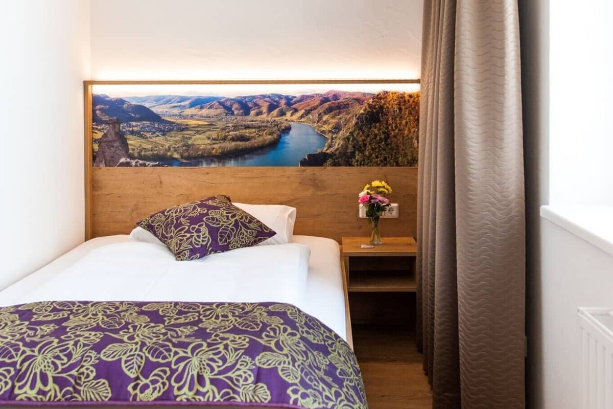 Une chambre à coucher avec une tête de lit en bois, un lit recouvert d’une courtepointe à motifs floraux violets, une table de chevet ornée d’un vase de fleurs et, au-dessus du lit, un grand tableau représentant un paysage.